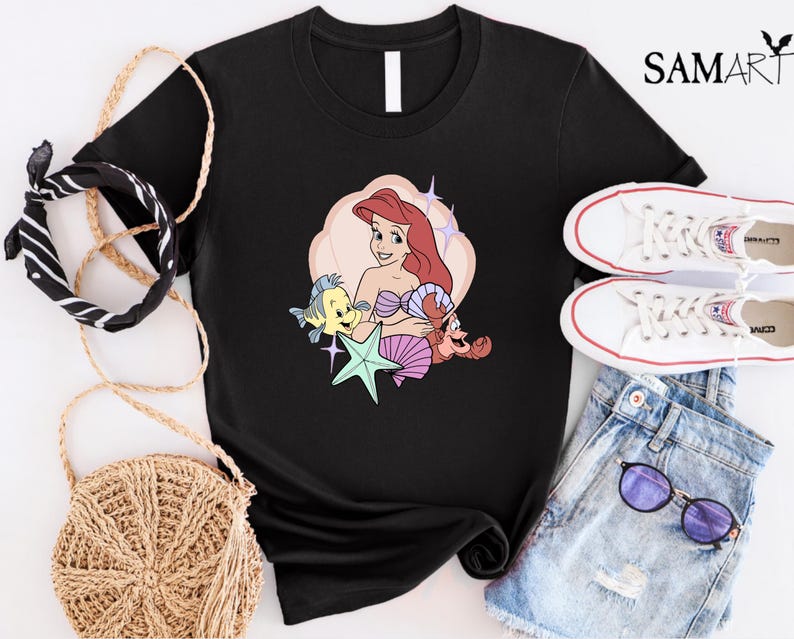 May include: Black t-shirt featuring a colorful graphic of Ariel from The Little Mermaid, with Flounder, Sebastian, and a starfish. The design is set against a light pink and cream background. The shirt is styled with white sneakers and denim shorts.