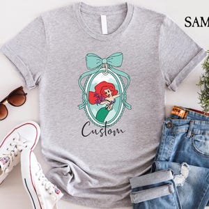 May include: A light gray t-shirt featuring a cartoon image of a mermaid with red hair and a green tail, framed by a teal bow and oval. The word "Custom" is written below the image. Sunglasses, white sneakers, and jeans are also in the image.