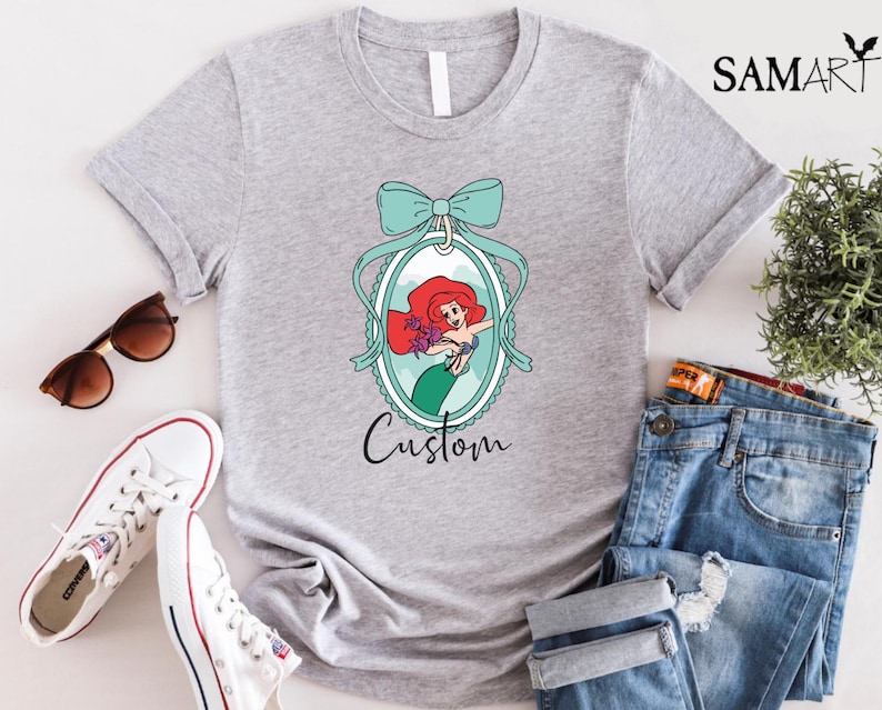 May include: A light gray t-shirt featuring a cartoon image of a mermaid with red hair and a green tail, framed by a teal bow and oval. The word "Custom" is written below the image. Sunglasses, white sneakers, and jeans are also in the image.