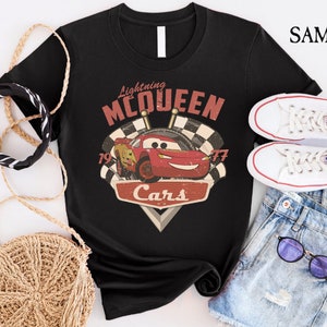 Op de afbeelding: Zwart T-shirt met een vintage-stijl afbeelding van Lightning McQueen uit de Disney Pixar film Cars. De afbeelding bevat de tekst "Lightning McQueen" en "Cars" met de nummers "19" en "77" aan weerszijden van de auto.
