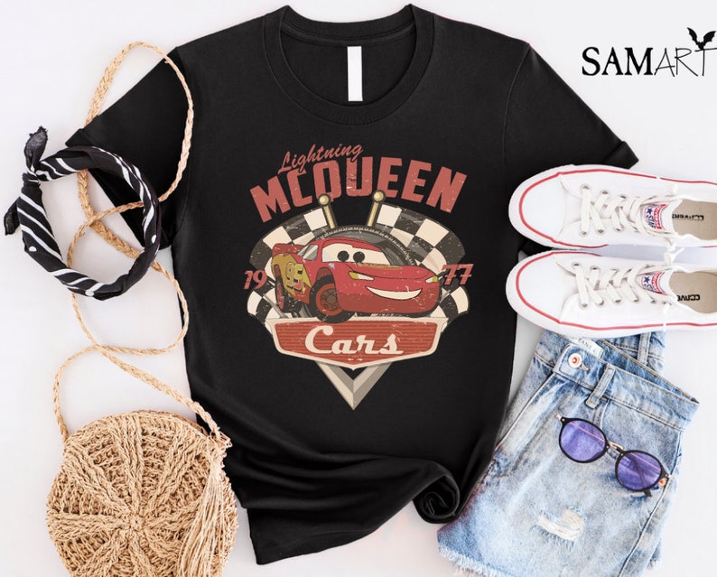 Op de afbeelding: Zwart T-shirt met een vintage-stijl afbeelding van Lightning McQueen uit de Disney Pixar film Cars. De afbeelding bevat de tekst "Lightning McQueen" en "Cars" met de nummers "19" en "77" aan weerszijden van de auto.