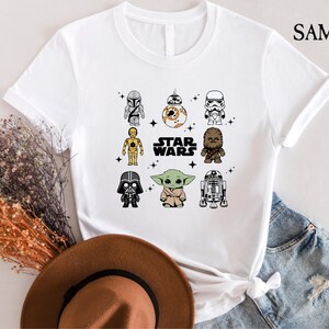 K&ouml;nnte beinhalten: Wei&szlig;es T-Shirt mit einem Star Wars-Design, das Cartoon-Charaktere wie Darth Vader, Yoda und R2-D2 zeigt. Der Text "STAR WARS" ist ebenfalls zu sehen. Das T-Shirt wird mit einem braunen Hut und Jeansshorts pr&auml;sentiert.