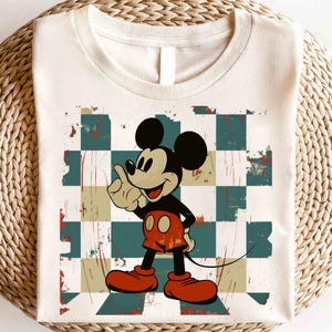 May include: A white t-shirt with a vintage-style graphic of Mickey Mouse on a checkered background. The graphic has a distressed look with faded colors and a worn texture.