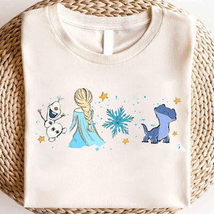 May include: Cream-colored t-shirt featuring a cartoon design with characters from a popular animated movie. The design includes a snowman, a princess in a blue dress, a snowflake, and a blue lizard-like creature, with yellow stars and blue dots.