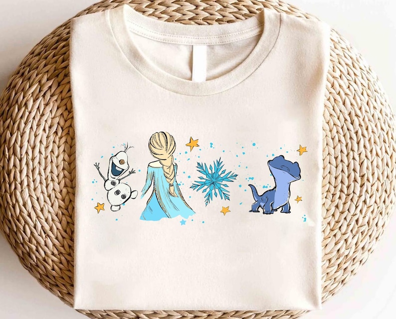 May include: Cream-colored t-shirt featuring a cartoon design with characters from a popular animated movie. The design includes a snowman, a princess in a blue dress, a snowflake, and a blue lizard-like creature, with yellow stars and blue dots.