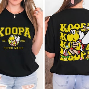 May include: Black t-shirt with a yellow and red Koopa Troopa graphic from Super Mario. The front design features the word "KOOPA" in yellow collegiate lettering, with "EST. 1985" and "SUPER MARIO" below. The back has a repeating Koopa design.