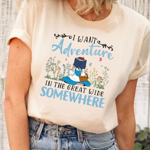 May include: A white t-shirt with a graphic of a girl reading a book in a field of flowers. The text on the shirt reads "I want adventure in the great wide somewhere."