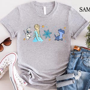 May include: A light gray t-shirt featuring a cartoon design of Elsa from Frozen, Olaf the snowman, a blue salamander, and a snowflake. The shirt also has the word "SAMART" printed on the upper right side.
