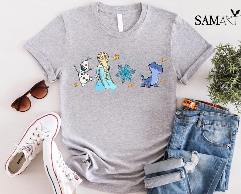 May include: A light gray t-shirt featuring a cartoon design of Elsa from Frozen, Olaf the snowman, a blue salamander, and a snowflake. The shirt also has the word "SAMART" printed on the upper right side.