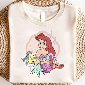 May include: Off-white t-shirt featuring a cartoon illustration of a mermaid with red hair, a purple seashell bra, and a smiling expression. A yellow fish, a red crab, and a green starfish are also depicted. The design is set against a pink heart-shaped background.