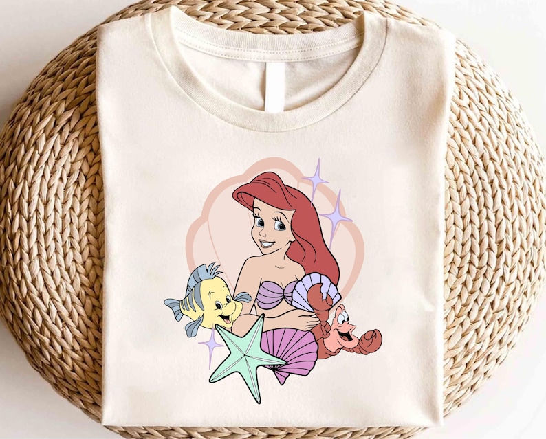 May include: Off-white t-shirt featuring a cartoon illustration of a mermaid with red hair, a purple seashell bra, and a smiling expression. A yellow fish, a red crab, and a green starfish are also depicted. The design is set against a pink heart-shaped background.