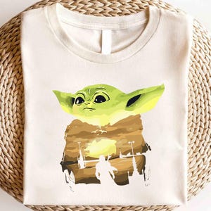 May include: Off-white t-shirt with a green cartoon character design, large ears, and a brown cloak. The graphic includes a landscape silhouette with a figure and structures. The shirt is made of a soft material.