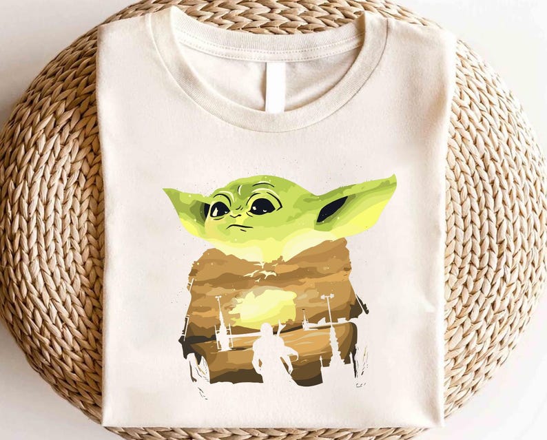 May include: Off-white t-shirt with a green cartoon character design, large ears, and a brown cloak. The graphic includes a landscape silhouette with a figure and structures. The shirt is made of a soft material.