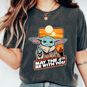 May include: A dark gray t-shirt with a graphic of a baby Yoda character from Star Wars. The graphic includes the text "Star Wars Day May the 4th Be With You".