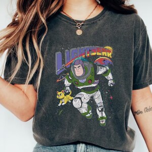 May include: A dark grey t-shirt featuring a graphic print of Buzz Lightyear from the Toy Story franchise. The graphic depicts Buzz Lightyear in a space suit with a yellow cat beside him. The text "LIGHTYEAR" is printed above the graphic.