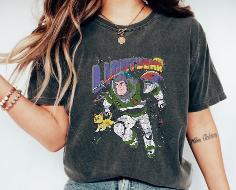 May include: A dark grey t-shirt featuring a graphic print of Buzz Lightyear from the Toy Story franchise. The graphic depicts Buzz Lightyear in a space suit with a yellow cat beside him. The text "LIGHTYEAR" is printed above the graphic.