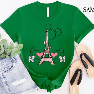 Puede incluir: Camiseta verde con una Torre Eiffel rosa y manos de Mickey Mouse sosteniendo corazones. La Torre Eiffel tiene una cabeza de Mickey Mouse rosa en la parte superior.