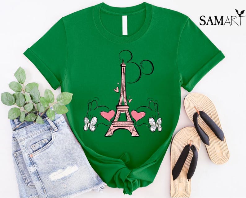Puede incluir: Camiseta verde con una Torre Eiffel rosa y manos de Mickey Mouse sosteniendo corazones. La Torre Eiffel tiene una cabeza de Mickey Mouse rosa en la parte superior.