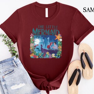 Puede incluir: Camiseta burdeos con una colorida ilustraci&oacute;n de "La Sirenita". El dise&ntilde;o incluye a Ariel y Eric en un bote, rodeados de plantas acu&aacute;ticas, una estrella de mar y el texto "The Little Mermaid."