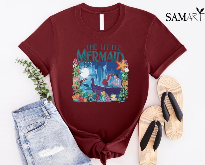 Puede incluir: Camiseta burdeos con una colorida ilustraci&oacute;n de "La Sirenita". El dise&ntilde;o incluye a Ariel y Eric en un bote, rodeados de plantas acu&aacute;ticas, una estrella de mar y el texto "The Little Mermaid."