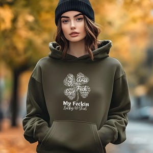 May include: A dark green hoodie with a white four-leaf clover graphic. The clover is made up of words, including "Feck" and "Arse". The text "My Feckin Lucky Shirt" is below the clover.