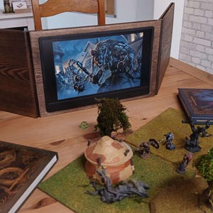 Może przedstawiać: Drewniany ekran do gier stołowych z cyfrowym wyświetlaczem przedstawiającym scenę bitwy fantasy. Ekran otoczony jest książkami, miniaturowymi figurkami i elementami terenu, tworząc konfigurację do gry fabularnej.