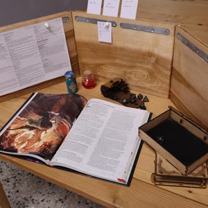 Może przedstawiać: Drewniany zestaw do gier stołowych z trójstronnym ekranem, książką, kostkami do gry i małym pudełkiem. Ekran ma biały papier z tekstem, a pudełko ma czarne filcowe wnętrze.