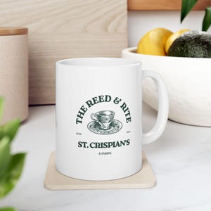 May include: White ceramic mug with a curved handle, featuring a green and white logo that reads "The Reed & Rite" and "St. Crispian's London." The logo includes a detailed teacup design. The mug sits on a beige coaster.