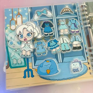 Puede incluir: Un libro de vestir azul y blanco con una variedad de pegatinas que representan a una ni&ntilde;a con cabello blanco, un ping&uuml;ino azul y una mochila azul. Las pegatinas se pueden usar para vestir a la ni&ntilde;a en el libro.