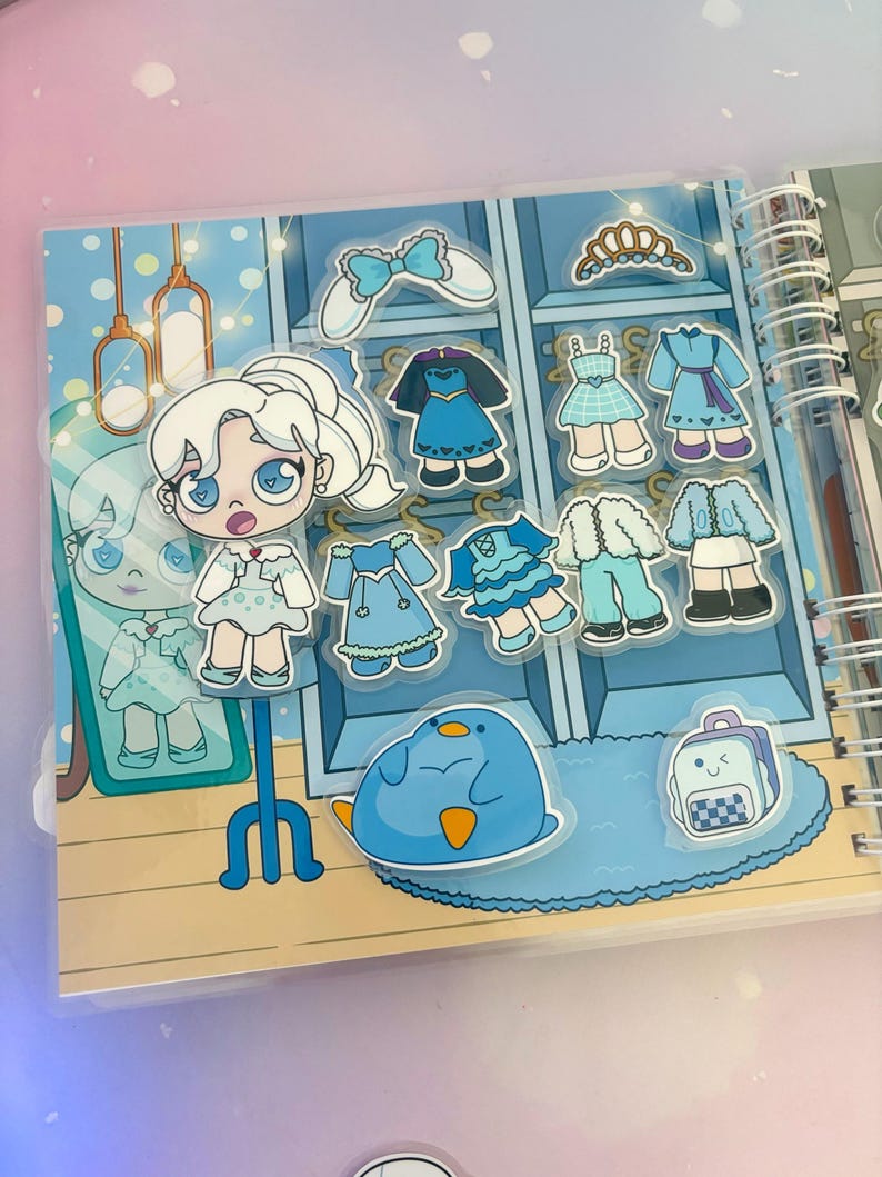 Puede incluir: Un libro de vestir azul y blanco con una variedad de pegatinas que representan a una ni&ntilde;a con cabello blanco, un ping&uuml;ino azul y una mochila azul. Las pegatinas se pueden usar para vestir a la ni&ntilde;a en el libro.