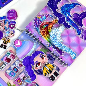 Puede incluir: Un cuaderno de espiral con una cubierta geométrica de color morado vibrante decorada con varios dijes acrílicos, incluyendo un personaje de dibujos animados con trenzas moradas, extensiones de pelo coloridas, productos de belleza y tijeras.