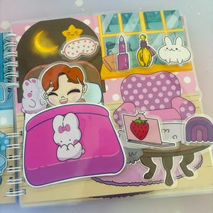 Puede incluir: Un libro ilustrado rosa y blanco con un personaje durmiendo en una cama rosa con un conejito blanco. El libro tiene una encuadernaci&oacute;n en espiral y una cubierta de pl&aacute;stico transparente. La ilustraci&oacute;n incluye un cielo nocturno con una luna y una estrella, una m&aacute;scara para los ojos rosa, una silla rosa con lunares blancos, una computadora port&aacute;til con una fresa en la pantalla, un arco&iacute;ris morado y blanco, y un conejito blanco con orejas rosas.