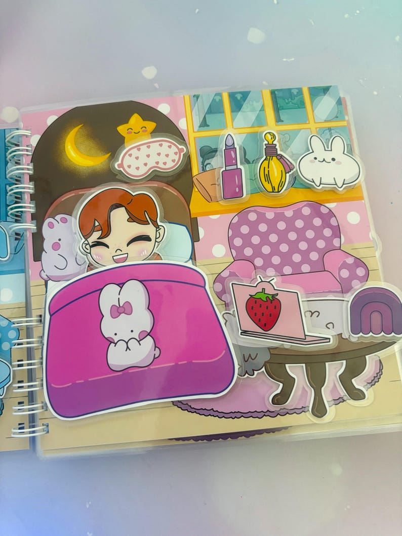 Puede incluir: Un libro ilustrado rosa y blanco con un personaje durmiendo en una cama rosa con un conejito blanco. El libro tiene una encuadernaci&oacute;n en espiral y una cubierta de pl&aacute;stico transparente. La ilustraci&oacute;n incluye un cielo nocturno con una luna y una estrella, una m&aacute;scara para los ojos rosa, una silla rosa con lunares blancos, una computadora port&aacute;til con una fresa en la pantalla, un arco&iacute;ris morado y blanco, y un conejito blanco con orejas rosas.