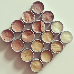 Puede incluir: Una colección de 14 pequeñas latas redondas de metal llenas de bálsamos labiales de diferentes colores. Las latas están dispuestas en forma de diamante sobre una superficie blanca.