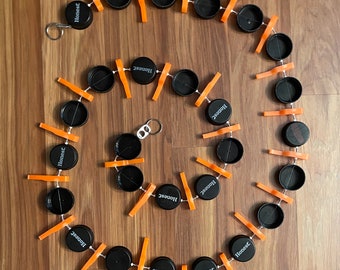 May include: A spiral chain made of black plastic bottle caps with the word "Honest" printed on them. The caps are connected by orange plastic clips and white string.