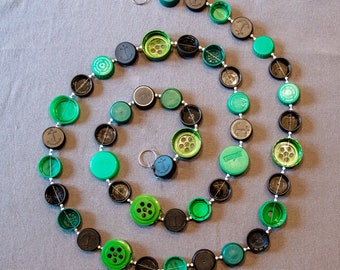 May include: A green and black necklace made from bottle caps with an anchor design. The necklace is strung together with silver beads and is in the shape of a spiral.