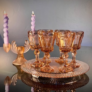 May include: A set of six amber-colored vintage goblets with ornate detailing, displayed on a matching glass serving tray. Two lit lavender-colored spiral candles and a gold-toned candle holder add to the elegant aesthetic.