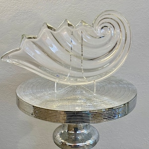 May include: A clear glass seashell-shaped bowl with a spiral design. The bowl is on a silver mirrored pedestal table.