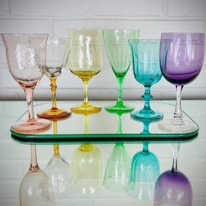 May include: A collection of six colorful glass goblets arranged on a rectangular mirror. The set includes a pale pink, a clear yellow-stemmed, a yellow, a bright green, a turquoise, and a deep purple glass, each featuring a unique stem design.