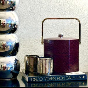 May include: A silver, multi-tiered lamp with a reflective surface sits on a black surface next to a burgundy ice bucket with a gold handle. Two gold-rimmed glasses and a book titled "Disco Years Ron Galella" are also on the surface.