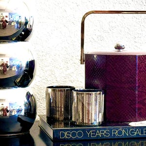 May include: A purple faux snakeskin ice bucket with a gold handle, two silver metallic glasses, and a book titled "Disco Years Ron Galella" on a black surface.