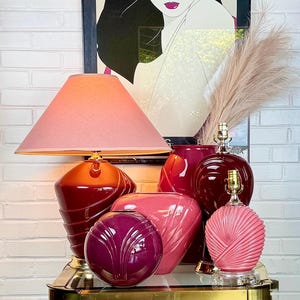 May include: A collection of ceramic decor items including a table lamp with a pink shade, a dark red spiral lamp base, a deep purple spherical vase, a pink fan-shaped vase, a tall dark red vase, and a small pink ribbed lamp base with gold accents.