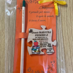 May include: An orange card with a wooden puzzle piece and pencil, tied with a yellow ribbon. The puzzle piece has text and a stack of books with an apple on top. The pencil has text that reads "Grazie per esserci".