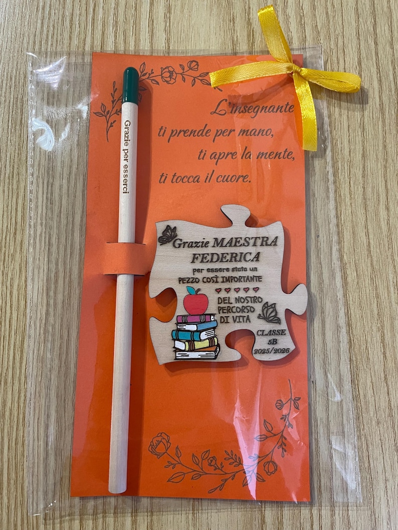 May include: An orange card with a wooden puzzle piece and pencil, tied with a yellow ribbon. The puzzle piece has text and a stack of books with an apple on top. The pencil has text that reads "Grazie per esserci".