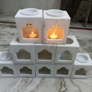 Könnte beinhalten: Ein Stapel weißer Marmor-Kerzenhalter mit aufwendigen Designs. Die Halter haben eine kleine Öffnung oben für ein Teelicht und eine größere Öffnung vorne mit einem dekorativen Bogenmuster.