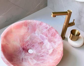 Pode incluir: Uma pia de pedra de quartzo rosa com uma torneira dourada. A pia redonda apresenta um padrão texturizado único e um orifício de drenagem. Uma pequena xícara dourada e uma esponja branca estão ao lado da torneira. Os acessórios do banheiro estão em um fundo neutro.