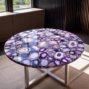 May include: Round coffee table with a polished, mosaic-like surface of purple and white agate stones. The table has a white metal base with an X-shaped design. The table is set on a neutral-colored rug.
