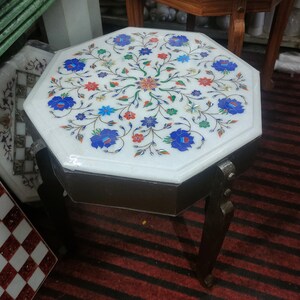May include: Octagonal white marble table with intricate floral inlay in blue, red, green, and gold. The table has a dark wood base with four legs.