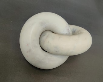 May include: White marble sculpture of two interlocking rings. The sculpture is smooth and has a subtle speckled pattern.