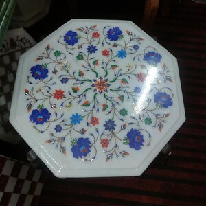 May include: Octagonal white marble table top with intricate floral design inlaid with colorful stones. The table top has a polished finish and is surrounded by a wooden frame.